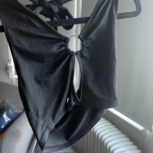 Svart bikini med ring och knyt - Snygg svart tröja med cool ringdetalj framtill och knytband i nacken. Tröjan har en stilren design med ett litet urklipp under ringen som ger en trendig touch. Materialet känns mjukt och följsamt mot huden.