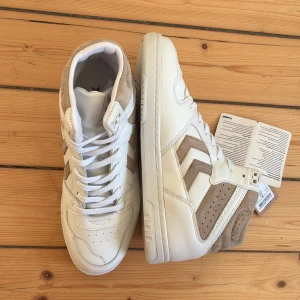 Vita och beige sneakers från Hummel X Design by Si - Snygga high-top sneakers från Hummel i vitt skinn och beige mocka. Klassisk siluett med perforerade detaljer och ikoniska Hummel-pilar på sidorna. Skorna har snörning och en platt sula. Storlek 40. Helt nya och oanvända. Etiketten sitter kvar.