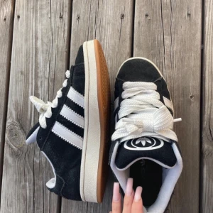 Adidas Campus sneakers svart/vit mocka - Svarta Adidas Campus sneakers i mocka med klassiska vita ränder på sidorna och vit Adidas-logga på plösen och hälen. Skorna har vit sula med gummidetalj och breda vita snören. Perfekta för dig som gillar retrostil och streetwear.