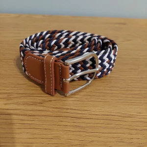 Randigt flätat bälte i blått och brunt - Snyggt flätat bälte med randigt mönster i marinblått, brunt och vitt. Bältet har detaljer i brunt skinn och ett klassiskt silverfärgat spänne. Perfekt accessoar för att lyfta din outfit med en sportig och stilren touch.