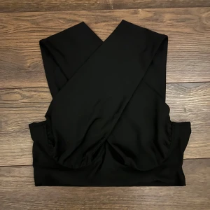 Svart omlott korsett-topp - Snygg svart korsett-topp med breda axelband som korsas framtill och omlott-detalj. Toppen är croppad och har en stilren, minimalistisk look. Perfekt för att lyfta din outfit med en edgy vibe. Tillverkad i mjukt, stretchigt material för skön passform.