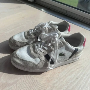 Lacoste T-Clip vita sneakers strl 44 - Snygga Lacoste T-Clip sneakers i vitt skinn och mocka med grå och marinblå detaljer samt röd häl. Klassisk krokodillogga på sidan och cool retrovibe. Skorna har snörning och en platt gummisula med Lacoste-mönster undertill. Perfekta för dig som gillar stilrena och sportiga sneakers.