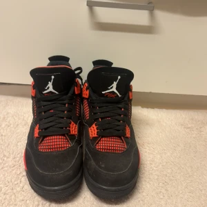Nike Air Jordan 4 svart/röd sneakers - Säljer ett par Nike Air Jordan 4 sneakers i svart och rött med klassisk Jumpman-logga på plösen och hälen. Skorna har meshpaneler, svarta snören och röda detaljer på sula och snörningsfästen. Ovandelen är i mocka och mesh för en sportig vibe.
