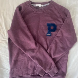 Lila sweatshirt från Panos Emporio - Säljer en lila sweatshirt från Panos Emporio med en stor blå P på bröstet. Tröjan har rund halsringning och långa ärmar. Perfekt för dig som gillar en avslappnad och stilren look.