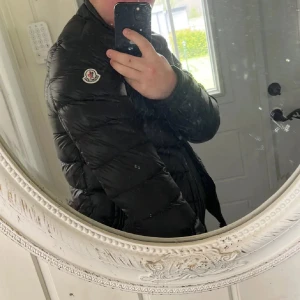 Svart dunjacka från Moncler, strl 3 - Säljer en svart Moncler dunjacka med klassisk logga på ärmen. Jackan är quiltad och har dragkedja framtill. Tillverkad i glansig polyester och fylld med dun, perfekt för kyliga dagar. Snygg och tidlös modell som passar till det mesta. Storlek 3 m/l