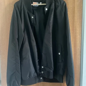 Svart bomberjacka med tryckknappar - Svart bomberjacka från Fruit of the Loom med klassisk ribbad mudd i midja och ärmslut. Jackan har tryckknappar framtill och två fickor med knappstängning. Enkel och stilren design, perfekt för dig som gillar en clean look.