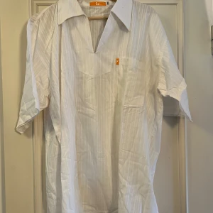 Vit randig henley från OZ, XXL - Vit henley-skjorta från O2 i storlek XXL. Skjortan har diskreta vertikala ränder, kort ärm, bröstficka och en öppen v-ringad krage. Tillverkad i lätt och luftig bomull, perfekt för varma dagar. Klassisk och stilren look med relaxed fit.
