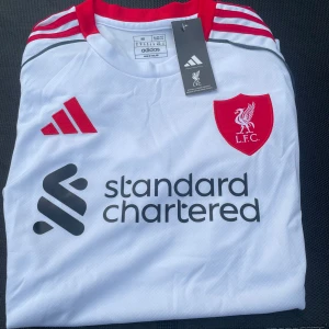 Liverpool vit bortatröja - Liverpool FC:s officiella bortatröja från Adidas i vitt med röda detaljer och klubbmärke på bröstet. Tröjan har korta ärmar, röd krage och tre röda ränder på axlarna. Tillverkad i lätt och ventilerande funktionsmaterial. Sponsorlogga på framsidan. Storlek M, helt ny med prislapp! frakt sker samma dag som köp, bättre pris än vad jag har hittar du knappast hör av dig vid fundering!