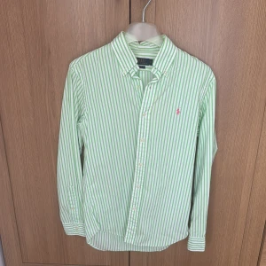 Grön- och vitrandig skjorta Polo Ralph Lauren - Snygg grön- och vitrandig skjorta från Polo Ralph Lauren i slim fit. Skjortan har långa ärmar, klassisk krage och knäppning framtill. Broderad rosa logga på bröstet. Perfekt för dig som gillar stilrena och fräscha plagg.