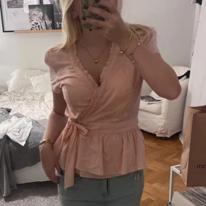 Rosa omlottblus med volang från Lots of Love - Supersöt ljusrosa omlottblus från Lots of Love med knyt i midjan och volangdetaljer längs v-ringningen. Blusen har korta ärmar och är gjord i mjuk bomull. Fint skick endast lite skrynklig 🩷