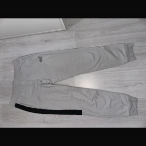 Grå EA7 mjukisbyxor med svart detalj - Grå mjukisbyxor från EA7 Emporio Armani med svart logotypband längs ena benet. Byxorna har resår i midjan och vid bensluten samt EA7-logga tryckt på höften. Perfekta för en chill och sportig look.