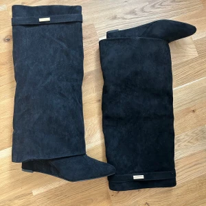 Svarta mockastövlar med klack och spetsig tå - Snygga svarta stövlar i mocka med spetsig tå och klack. Stövlarna har en stilren design med en diskret gulddetalj upptill. Perfekta att styla med kjol eller klänning för en trendig look. Storlek 40 men passar 39. Klacken är 7cm. Endast testade.