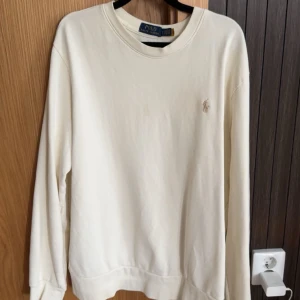 Vit sweatshirt från Polo Ralph Lauren - Klassisk vit sweatshirt från Polo Ralph Lauren med rund halsringning och broderad logga på bröstet. Tröjan har lång ärm och är tillverkad i mjuk bomull, perfekt för en clean och stilren look.