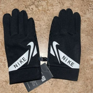 Svarta Nike träningshandskar - Svarta träningshandskar från Nike med vit logga och text på ovansidan. Handflatan har greppvänliga mönster i svart och fingertopparna är anpassade för touchskärm. Snygg, sportig design som passar perfekt till utomhusträning.