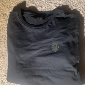 Svart långärmad tröja från EA7 - Snygg svart långärmad tröja från EA7 Emporio Armani med rund halsringning och diskret EA7-logga på bröstet. Tillverkad i mjuk bomull som känns skön mot huden. Perfekt för dig som gillar stilrena och enkla plagg med en exklusiv touch.