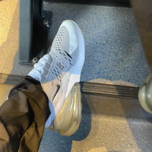 Nike Air Max 270 vita sneakers - Säljer ett par Nike Air Max 270 sneakers i vitt med mesh-överdel och stora genomskinliga Air Max-sulan. Skorna har snörning, diskret svart swoosh och 270-detalj på sidan. Perfekta för dig som gillar sportig och modern stil.