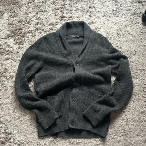 Mörkgrå kofta från Dressmann, M - Mysig mörkgrå kofta från Dressmann i ribbstickad ullmix. Koftan har sjalkrage, knäppning med  knappar framtill och två fickor. Perfekt för lager-på-lager och  höst- eller vinterstil.