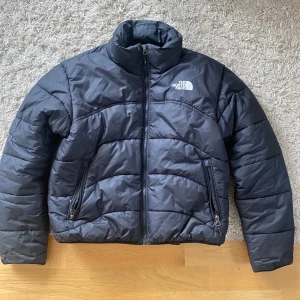Svart pufferjacka från The North Face - Svart pufferjacka från The North Face i klassisk modell med hög krage och två dragkedjeförsedda fickor. Jackan har quiltad design och logga på både bröst och rygg. Perfekt för kyliga dagar och enkel att matcha med det mesta.