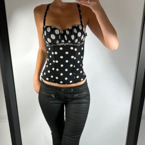 Svart prickig korsett-topp - Supersnygg svart korsett-topp med vita prickar och smala axelband. Toppen har en figurnära passform och rynkad byst med volangkant. Perfekt för dig som vill sticka ut med ett lekfullt mönster och trendig siluett.