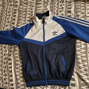 Adidas blå och vit vindjacka - Snygg Adidas vindjacka i blått, vitt och marinblått med klassiska tre ränder på ärmarna. Jackan har dragkedja, höga muddar och två fickor med blå detaljer. Stor Adidas-logga på ryggen och mindre logga på bröstet. Perfekt för en sportig streetstil.