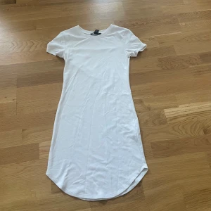 Vit kort t-shirtklänning H&M - Enkel och stilren vit t-shirtklänning från H&M med rund halsringning och korta ärmar. Klänningen har en rak passform och är tillverkad i mjuk bomull, perfekt för en avslappnad look. Längden är kort och nederkanten är lätt rundad.