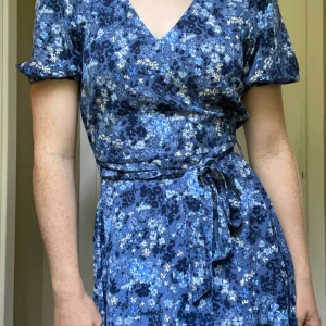 Blommig blå klänning med knyt - Söt längre blå klänning med blommigt mönster och knytning. Klänningen har korta, puffiga ärmar, v-ringning och knytband i midjan för en snygg passform. 