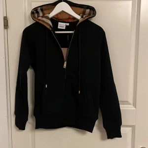 Svart Burberry hoodie  - Svart hoodie från Burberry med klassisk rutig huva i beige, rött och svart. Tröjan har dragkedja framtill, snörning vid huvan och diskret broderad logga på bröstet. Perfekt för dig som vill ha en stilren och lyxig streetwear-look. Nyskick och aldrig använd 