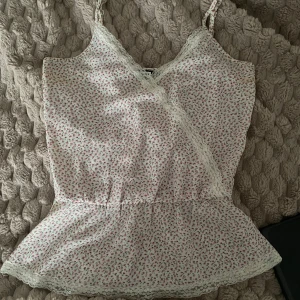 Blommig omlott linnetopp med spets - Supersöt vit linnetopp med små rosa blommor och tunna axelband. Toppen har omlott-skärning framtill och är dekorerad med vit spets längs kanterna. Den har en lätt utställd nederdel och är perfekt för sommarens varma dagar.