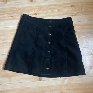 Svart kortkjol i mockaimitation från Divided - Svart A-linjeformad kortkjol från Divided med snygga silverfärgade knappar framtill. Kjolen är gjord i mjuk mockaimitation och har två fickor fram. Perfekt för dig som gillar en stilren och trendig look.