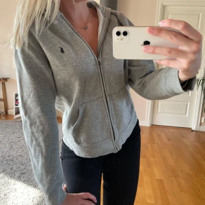 Grå hoodie från Ralph Lauren - Grå hoodie från Ralph Lauren med dragkedja framtill och klassisk huva. Tröjan har två fickor och den ikoniska lilla broderade loggan på bröstet. Perfekt för vardagen och enkel att matcha med allt. Mjuk och skön bomullskvalitet. Det är L i barnstorlek så skulle säga att den motsvarar xs/s!💗