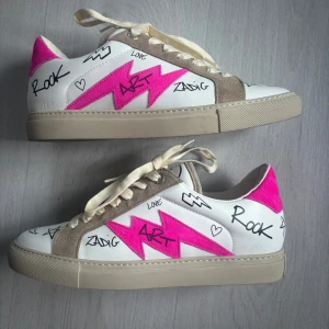 Zadig & Voltaire sneakers  - Unika sneakers från Zadig & Voltaire med vit bas, beige mockadetaljer och starkt rosa blixt på sidan. Skorna har coola graffiti-liknande texter och symboler i svart, samt snörning och platt sula. Perfekta för dig som vill sticka ut med din stil.