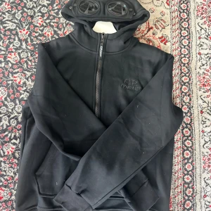  CP Company Hoodie - Svart hoodie från CP Company med ikonisk huva som har inbyggda goggles. Tröjan har dragkedja framtill, ribbade muddar och broderad logga på bröstet. Perfekt för dig som gillar streetwear och vill sticka ut med en unik detalj på huvan.