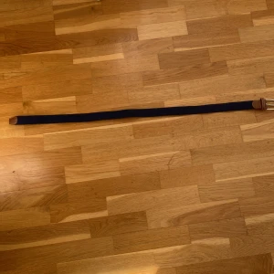 Mörkblå flätad bälte med skinn - Snyggt mörkblått flätat bälte med bruna skinn-detaljer och silvrigt spänne. Bältet har en stilren design med tydliga sömmar på skinnet och är gjort i ett mjukt syntetmaterial som är flexibelt och bekvämt. Perfekt för att ge din outfit en clean och modern touch.