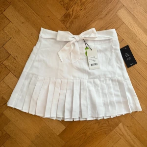 Vit plisserad kjol med rosett, EU 38 - Superfin vit kortkjol med breda plisserade veck och ett fint band i midjan. Kjolen har en klassisk och fräsch look, perfekt för dig som gillar stilrena detaljer. Kjolen är aldrig använd och har lappen kvar. Dock märkte jag av en liten fläck som jag inte testat att tvätta bort eftersom den inte är använd. Fläcken är inte mycket synlig och tror absolut den borde gå bort, men kan inte garantera något.