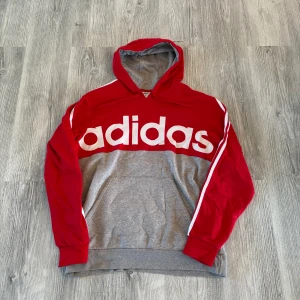 Röd och grå Adidas hoodie - Snygg hoodie från Adidas i rött och grått med stor vit logga över bröstet. Klassiska vita ränder längs ärmarna, huva och känguruficka framtill. Perfekt för en sportig och avslappnad stil.