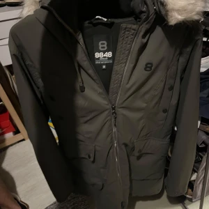 Parkasjacka 8848 Altitude med fuskpäls - Mörkgrön parkasjacka från 8848 Altitude med huva och snygg fuskpäls. Jackan har dragkedja, knappar och flera fickor framtill. Perfekt för kalla dagar med sin långa modell och stilrena look.