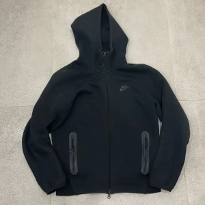 Svart Nike hoodie med dragkedja - Svart hoodie från Nike med hel dragkedja och två fickor med dragkedja framtill. Klassisk logga på bröstet och stor huva. Tillverkad i mjuk bomullsblandning, perfekt för chill eller träning. Snygg och enkel design som funkar året runt.