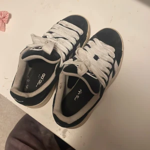 Adidas Campus sneakers svart/vit strl 36 - Säljer ett par klassiska Adidas Campus sneakers i svart mocka med vita detaljer och breda vita snören. Skorna har den ikoniska tre-randiga designen på sidorna och en beige sula. Perfekta för dig som gillar retrostil och streetwear.