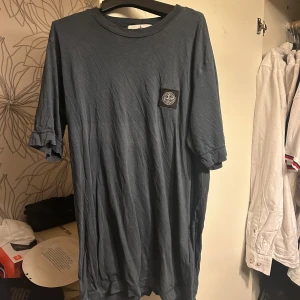 Blå t-shirt från Stone Island - Säljer en blå t-shirt från Stone Island med klassisk rund hals och korta ärmar. T-shirten har en patch med loggan på bröstet och är gjord i mjuk bomull. Perfekt för dig som gillar streetwear och vill ha något stilrent med ikonisk branding.