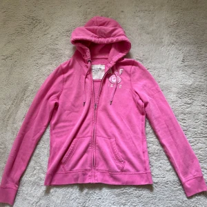 Rosa hoodie från Abercrombie & Fitch - Säljer en rosa hoodie från Abercrombie & Fitch med dragkedja och huva. Tröjan har broderad logga och text på bröstet, snörning i huvan och två fickor framtill. Perfekt för en chill och avslappnad stil.