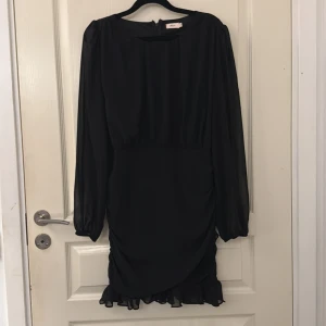 Svart omlottklänning med volang - Snygg svart kortklänning från Nelly  med långa ärmar i transparent tyg, omlottdesign och volangdetalj längst ner. Perfekt för dig som gillar stilrent 