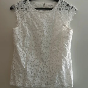 Vit spetsblus från Holly & Whyte - Superfin vit blus i spets från Holly & Whyte by Lindex. Blusen är ärmlös med rund halsringning och har ett vackert spetsmönster över hela plagget. Baksidan har en liten knappdetalj i nacken. Perfekt för dig som gillar romantisk och stilren look.