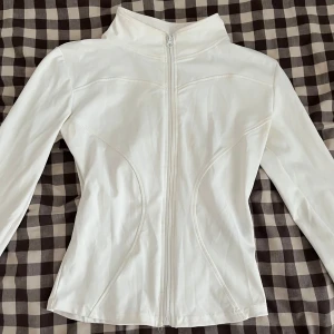 Vit slim fit jacka med dragkedja - Säljer en vit, figursydd tränings jacka med hel dragkedja framtill och hög krage. Jackan har långa ärmar och är tillverkad i ett stretchigt, slätt material som ger en sportig och clean look. Perfekt för dig som gillar stilrena och enkla plagg. Superfin på! 