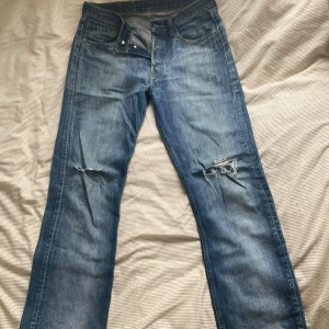 Levi's blå jeans med slitningar - Klassiska Levi's jeans i blå denim med snygga slitningar och hål på knäna. Rak passform, femficksmodell och knappgylf. 