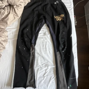 Svarta mjukisbyxor Gallery Dept. - Svarta mjukisbyxor från Gallery Dept. med gult tryck på ena benet, elastisk midja med snörning och unika färgstänk och penseldrag. Byxorna har sidofickor och paneler i grått längs benen. Tillverkade i 100% bomull för en skön och avslappnad känsla. Bra skick