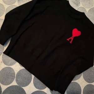 Svart stickad tröja från Ami Paris - Svart stickad tröja från Ami Paris med röd broderad logga i form av ett hjärta och bokstaven A på bröstet. Tröjan har rund halsringning, långa ärmar och breda ribbade muddar vid ärmslut och nederkant. Perfekt för en stilren look.
