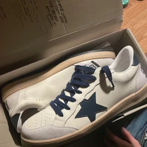 Golden Goose sneakers vit/blå stjärna - Golden Goose sneakers i vitt skinn med blå stjärndetalj på sidan och blå snörning. Yttersulan är beige och har en svart undersida med stjärnmönster. Tåpartiet har inslag av mocka och skon har en klassisk låg profil. Perfekt för dig som gillar stilrena men ändå unika sneakers.