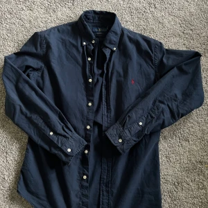 Mörkblå skjorta från Ralph Lauren - Klassisk mörkblå skjorta från Ralph Lauren med button-down krage och broderad röd logga på bröstet. Skjortan har långa ärmar, vita knappar och är tillverkad i mjuk bomull. Perfekt för en stilren och avslappnad look.