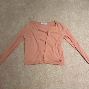 Ljusrosa kofta från H&M - Söt ljusrosa kofta från H&M i mjuk bomull. Koftan har långa ärmar, knappar framtill och en liten gulddetalj vid nederkanten. Perfekt att slänga över en topp för en chill look.