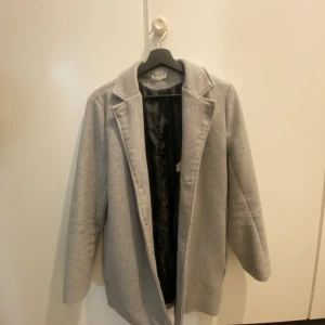 Ljusgrå kappa med klassisk krage - Snygg ljusgrå kappa med klassisk krage och rak passform. Jackan har helfodrad insida och långa ärmar, perfekt för kyligare dagar. Enkel och stilren design som passar till det mesta.